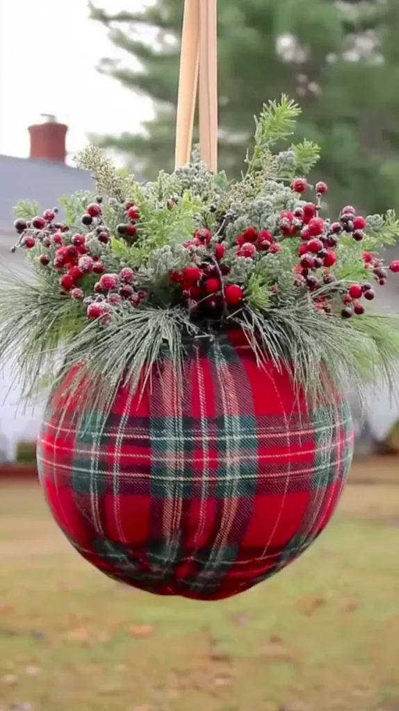 A giant Christmas ornament wrapped tightly in red and green plaid fabric, topped with realistic fir branches, frosted pine needles, red berries, and subtle winter greenery, hanging outdoors on a neutral ribbon, soft natural lighting, crisp textures, gentle shadowing, cozy holiday atmosphere