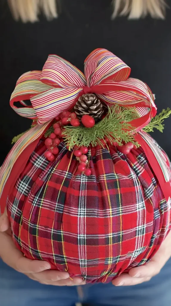 A large handmade Christmas ornament wrapped in red plaid fabric with crisp folds, topped with a large red bow, curled striped ribbon, pinecones, evergreen sprigs, and red berries, photographed indoors with soft holiday lighting, warm and festive mood 