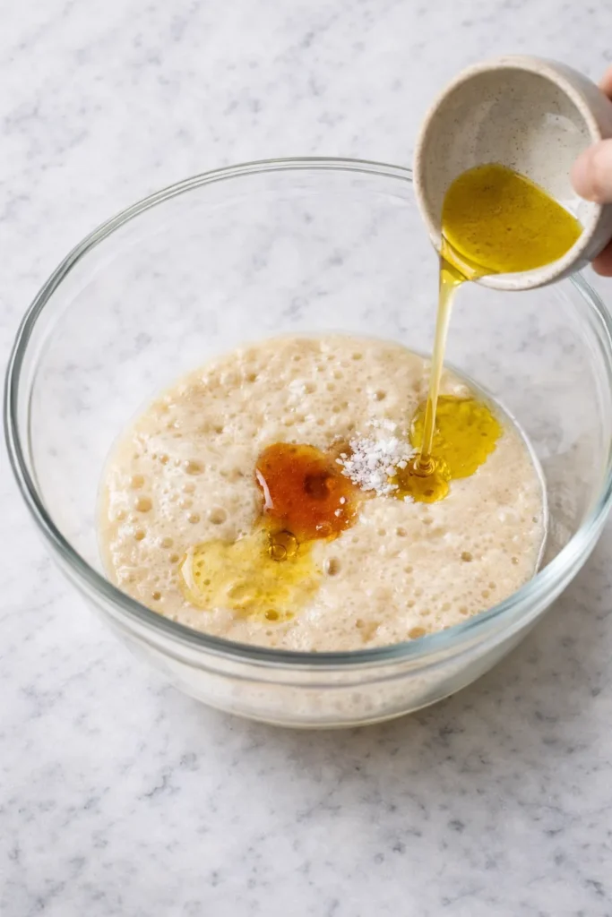 Olive oil poured into gluten-free sourdough starter with honey and salt in a glass bowl on white marble.