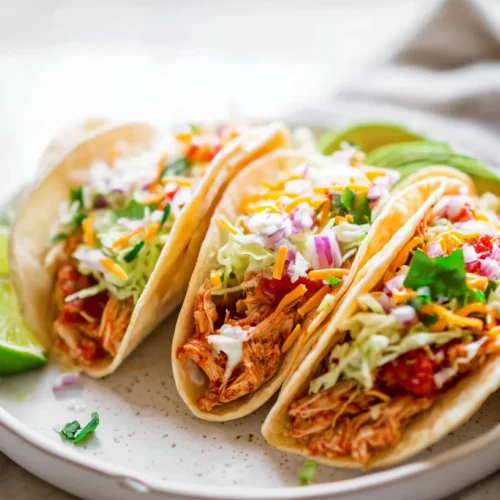 Three shredded chicken tacos on a white plate with lettuce, cheese, sour cream, red onion, cilantro, and lime wedges on a white marble counter.
