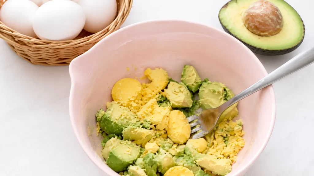 Light pink mixing bowl with avocado chunks and hard-boiled egg yolks for guacamole deviled eggs, with a fork, whole eggs in a basket, and a halved avocado nearby.
