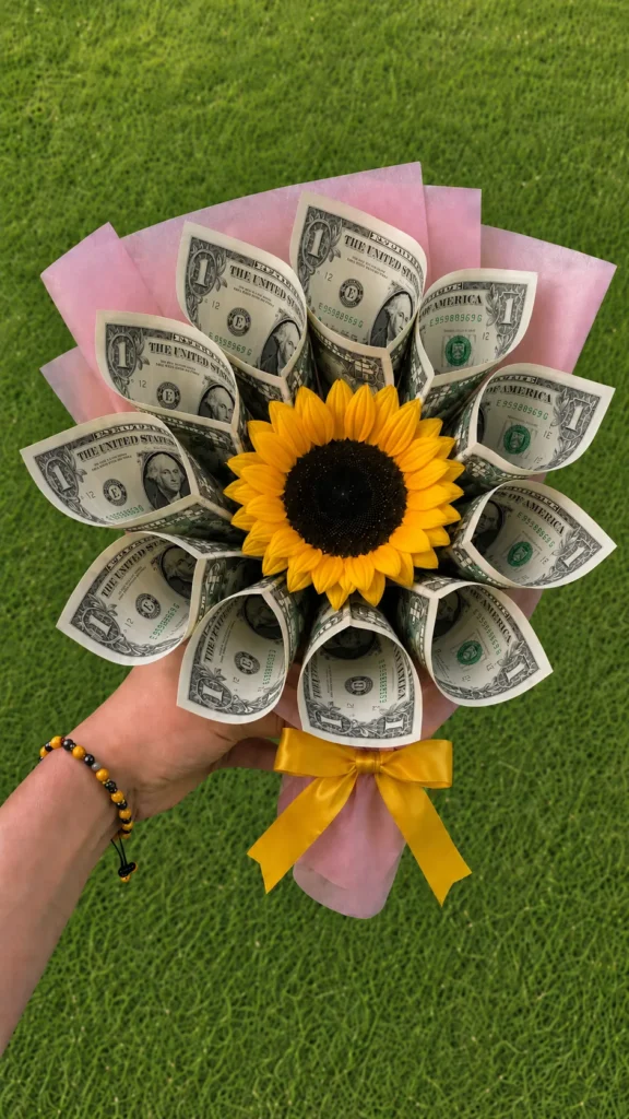 Small sunflower money bouquet with folded U.S. dollar bill petals, pink wrapping paper, yellow ribbon, and green grass background.