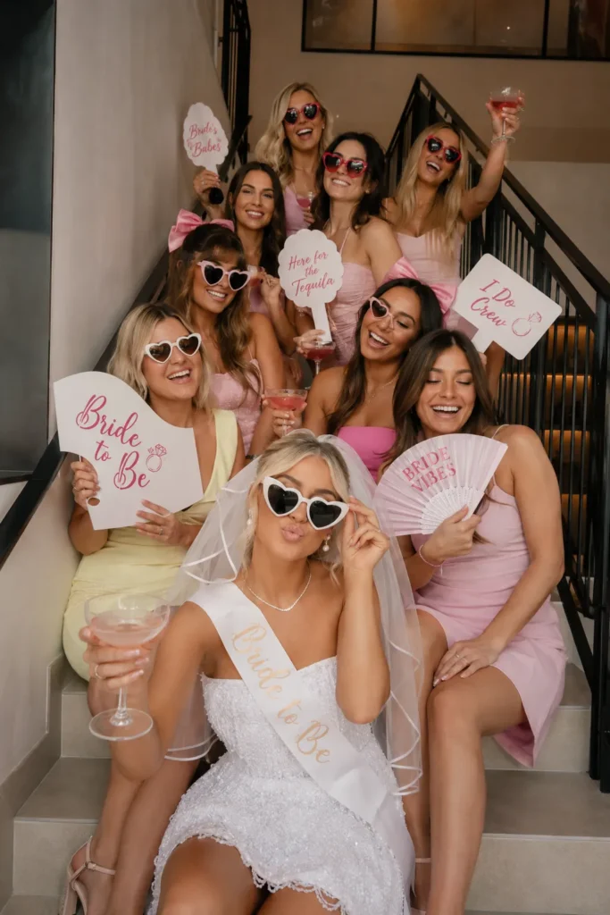 Group of women in pink and white outfits gathered on stairs for a bachelorette party, wearing sunglasses and holding fun props like "Bride to Be" signs and cocktails