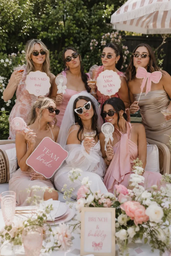 Bride and bridesmaids in pink and white attire holding custom photo props at an elegant outdoor brunch celebration under a striped umbrella with floral decorations