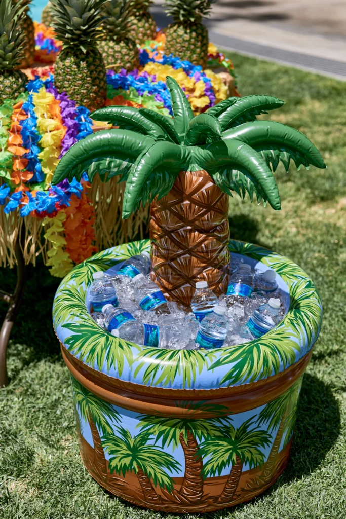 Inflatable palm tree cooler filled with ice and blue water bottles on green artificial turf, with colorful flower decorations in background