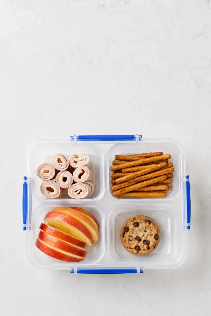 Divided lunch container with sliced apple, pretzel rolls, breadsticks, and chocolate chip cookie on marble surface