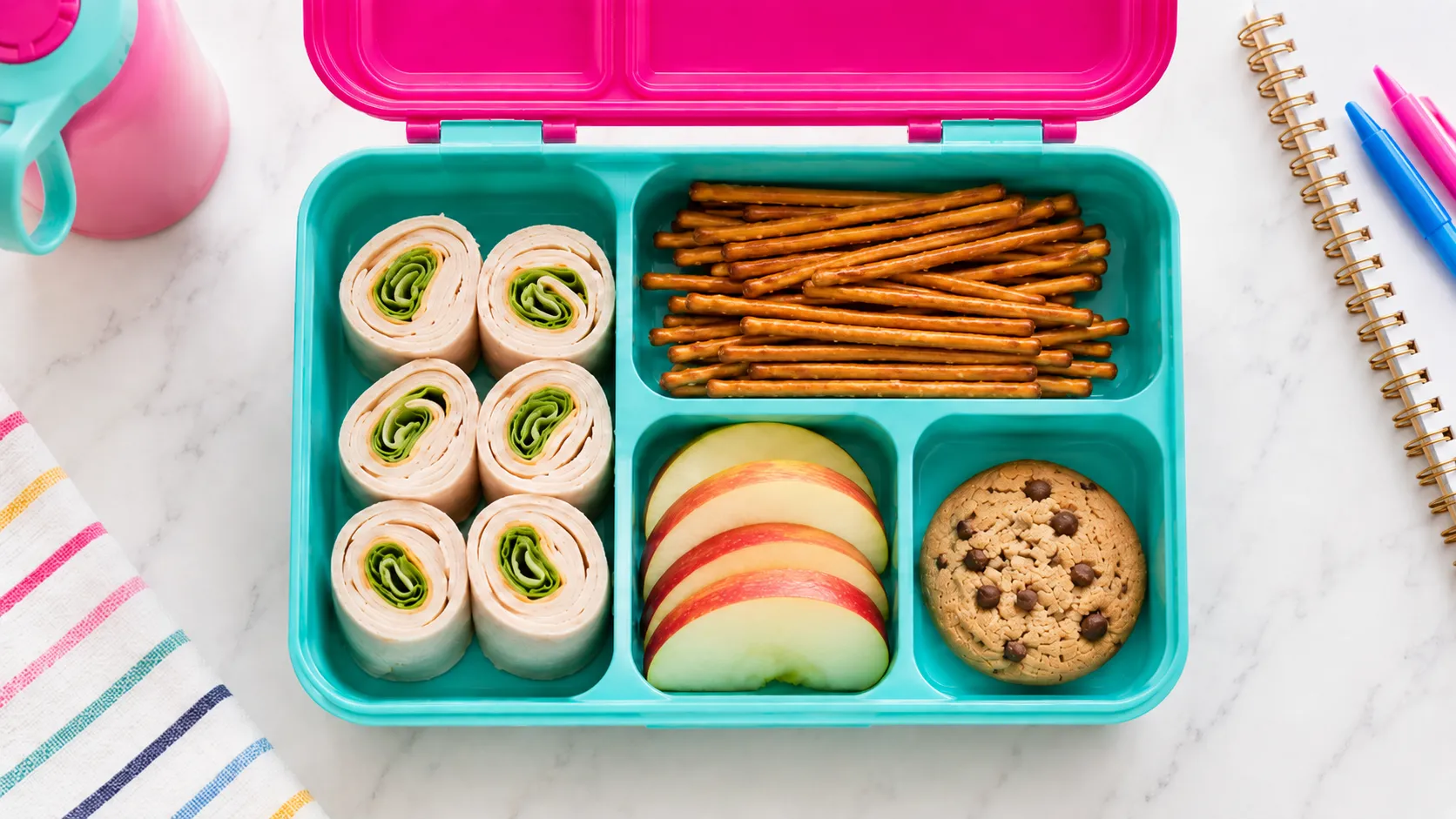 Turquoise bento box filled with pinwheel sandwiches, pretzel sticks, apple slices, and a chocolate chip cookie on marble surface with school supplies nearby