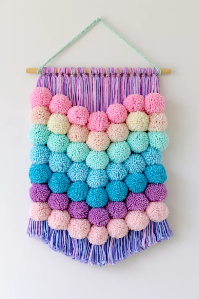Colorful handmade wall hanging with purple macramé top, rows of pink and cream pom poms in the middle, and turquoise and blue pom poms below, finished with purple fringe trim