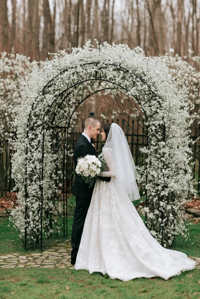 Bride and groom standing under a lush white flowering arch in a garden setting, with bare trees in the background