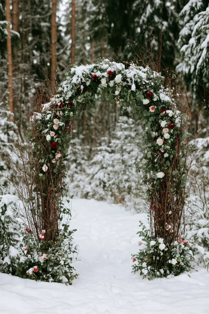 Rustic wedding arch adorned with red, pink, and white roses covered in fresh snow, set in a snowy evergreen forest