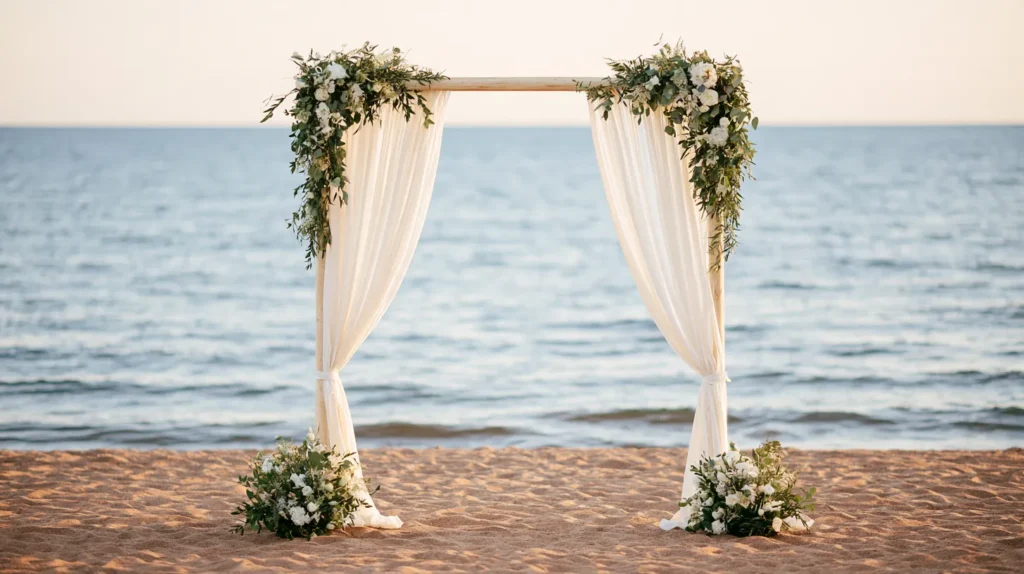 Wooden wedding arch with flowing cream drapes and greenery garland on sandy beach with ocean waves behind