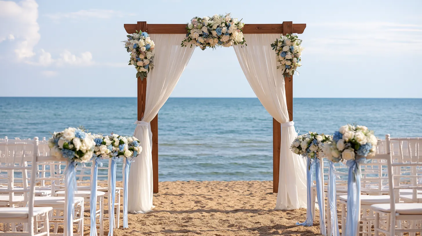 Wooden wedding arch with cream draping and white floral arrangements on a sandy beach with ocean backdrop and white chairs lining the aisle