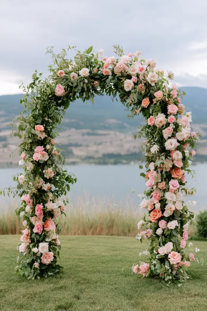 Romantic wedding arch adorned with pink and cream roses and green foliage, set in a grassy field with mountains and lake in the background