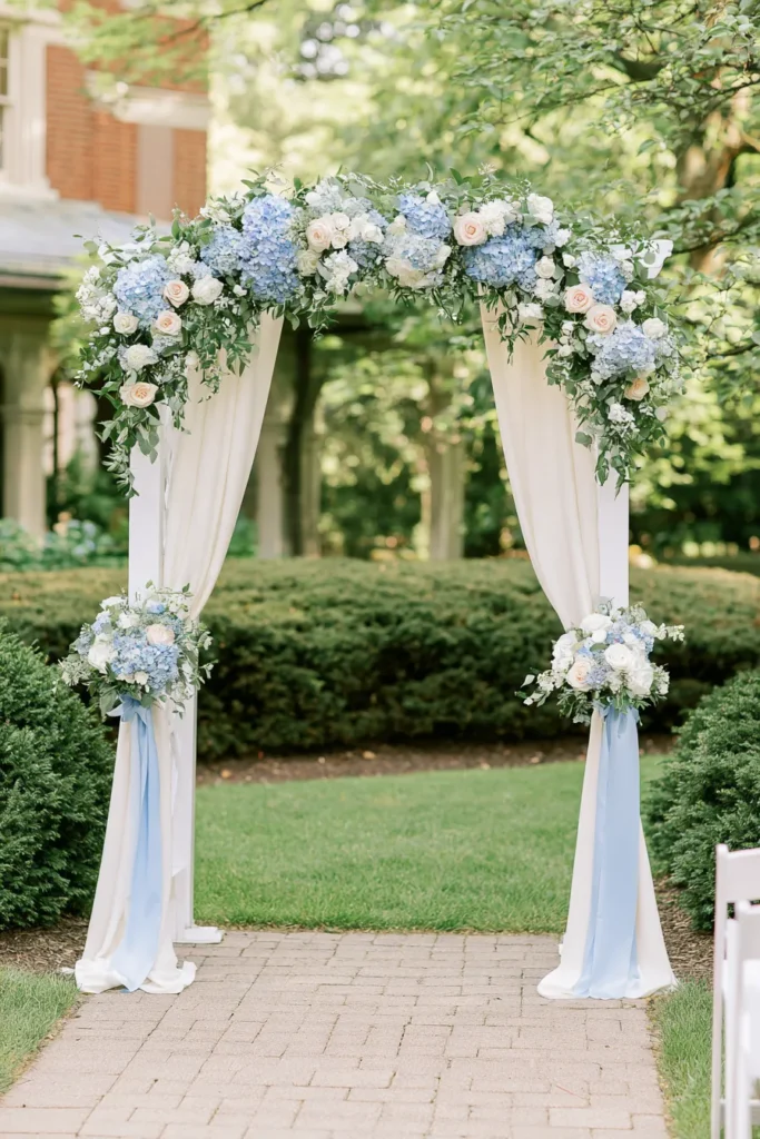 White wedding arch with blue hydrangeas, blush roses, and greenery draped in sheer fabric in a manicured garden setting
